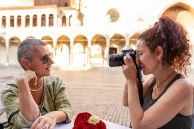 Orta yaşlı güzel bir çift, tarihi bir meydanda, açık hava masasında otururken fotoğraf çekilirken eğleniyorlar.