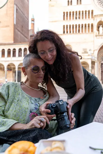 İki orta yaşlı bayan arkadaş, tarihi bir meydanda açık hava masasında kamera görüntülerine bakıyor.