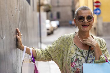 Tıraşlı saçlı, renkli alışveriş torbalarıyla siyah bir duvarın önünde poz veren zarif olgun bir kadının portresi.