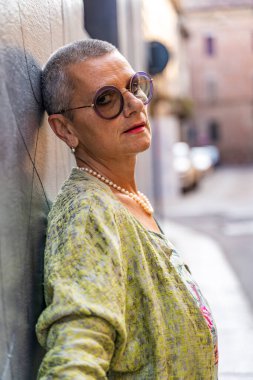 Sokakta siyah duvara yaslanmış kameraya bakan güzel olgun bir kadının portresi.