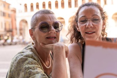 Birkaç orta yaşlı bayan arkadaş tarihi bir meydanda açık bir kafede oturmuş menüye bakıyorlar.