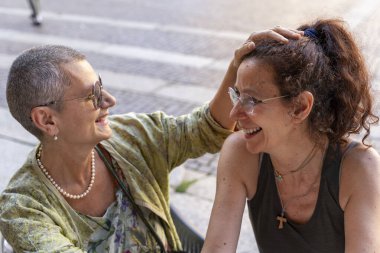 İki güzel orta yaşlı kadın, tarihi bir meydanda açık bir kahve barında karşılıklı şefkat göstererek eğleniyorlar.