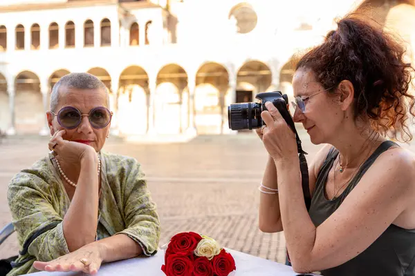 Orta yaşlı güzel bir çift, tarihi bir meydanda, açık hava masasında otururken fotoğraf çekilirken eğleniyorlar.