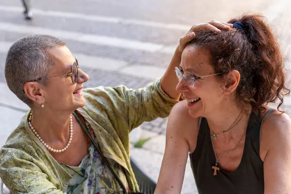 İki güzel orta yaşlı kadın, tarihi bir meydanda açık bir kahve barında karşılıklı şefkat göstererek eğleniyorlar.