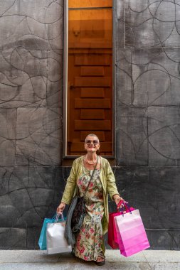 Zarif olgun bir kadın siyah bir duvarın önüne renkli alışveriş torbaları atarak eğleniyor.