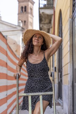 Güzel, orta yaşlı bir kadın şehir inşaatında hasır şapka takarak bir teslimat için el arabası itiyor.