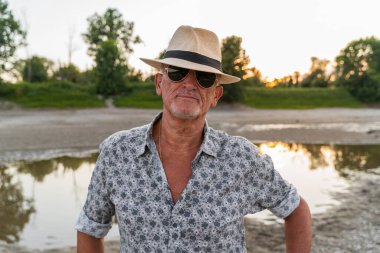 Gün batımında nehir kıyısında panama şapkası ve güneş gözlüğü takan orta yaşlı bir adamın portresi.