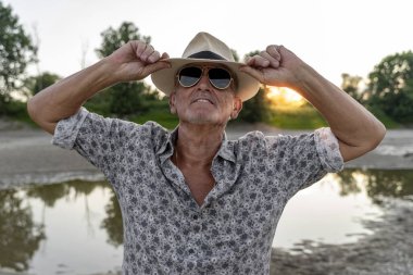 Gün batımında nehir kıyısında panama şapkası ve güneş gözlüğü takan orta yaşlı bir adamın portresi.