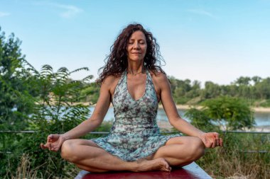 Orta yaşlı bir kadın yaz elbisesi giyiyor. Masada oturuyor. Gün batımında yoga pozisyonunda nehir kenarında dinleniyor.