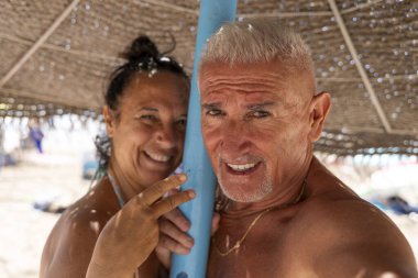 Mutlu ve güzel bir emekli çift yaz tatili sırasında kumsalda saman şemsiyesi altında selfie çekerken sarılıp gülümsüyor.