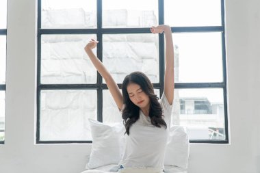 Healthy asian woman waking up relax muscles beside window early in morning. Happy young girl stretching raised arms muscles body cozy after waking up sitting on bed with pillow at apartment. Lifestyle