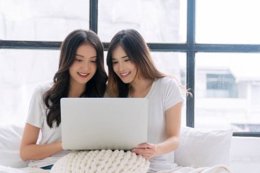 Cheerful asian LGBTQ lesbian woman using laptop online sitting together with pillows white bed at apartment. Happy two young girl couple leisure with notebook working online. Technology communication