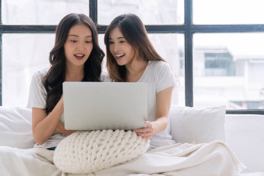 Cheerful asian LGBTQ lesbian woman using laptop online sitting together with pillows white bed at apartment. Happy two young girl couple leisure with notebook working online. Technology communication