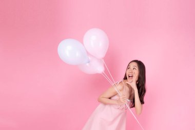 Excited cheerful asian woman holding balloons and hands beside mouth smiling with toothy standing over isolated pink background. Joyful teenager girl with pastel balloons shocked  amazed expression.