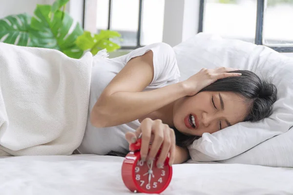 Upset asian young woman displeased stopping ringing red alarm clock on white bed in morning. Angry unhappy young girl sleeping with white pillow holding hand bell snooze alarm clock laziness at home.