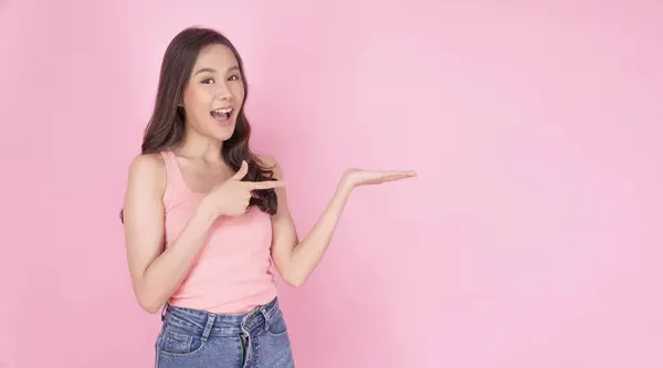 エキサイティングなアジアの女性は ピンクの背景に立っている空の指を使用して手を上げました 孤立した製品を提示する手のコピースペースを示す自信のある笑顔の若い少女 広告プレゼンテーション — ストック写真