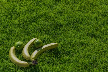 Bu fotoğraf gerçekçi bir şekilde çimenli bir çimenlikteki iki muz ve limonu gösteriyor. Muzlar çimlerin üzerinde yatıyor. Limon muzların yanında yatıyor. Çimler yeşil ve yeni kesilmiş gibi görünüyor.. 