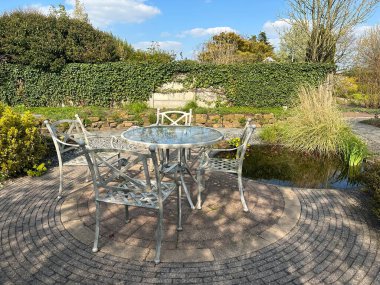 Avlu. Bahçe yemek takımı. Eski moda kıvrımlı demir teras sandalyeleri ve masa. Bahar manzarası: Taş teras, gölet, taş duvar, yeşillik. Başvuru, Hollanda, 09 Nisan 2023