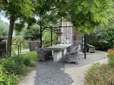 Apeltern, Hollanda, 11 Temmuz 2023, Appeltern macera bahçesi. Bahçe çardağı, cam yapı, beton masa ve rattan sandalyeler. Ağaç Tetradium daniellii (arı-arı ağacı)