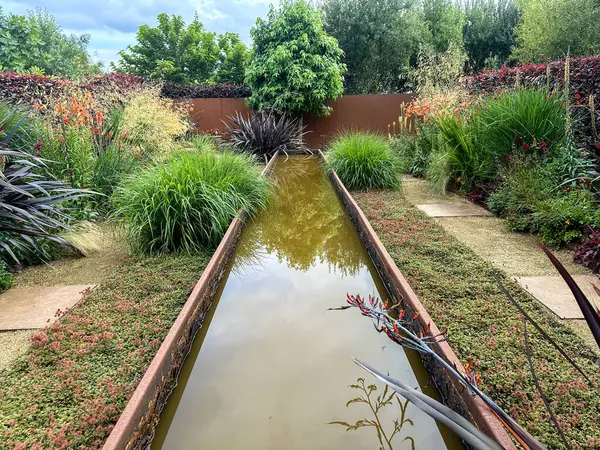 Phormium Black Adder ve Acaena Kupferteppich 'in zemin örtüsü de dahil olmak üzere canlı turuncu ve kırmızı bitkilerle çevrili paslı metal gölet. Kerfouler Bahçeleri, Fransa