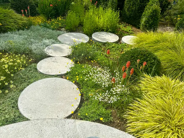 Taş kaldırım. Yuvarlak taşlı kıvrımlı yol yemyeşil bitkiler ve canlı çiçeklerle çevrilidir. Kerfouler Bahçeleri, Fransa