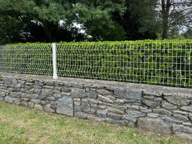 Kahverengi taştan duvar temeli üzerine kurulmuş beyaz çift halkalı tel örgülü tel örgü. Çit arka planda yemyeşil bir çit tarafından çerçevelenmiştir. Langourla, Le-Mene (Brittany, Fransa)