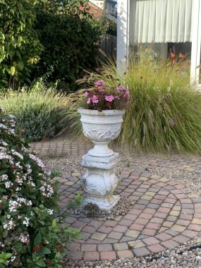 Süslü beyaz vazoda dairesel tuğla verandalı ve pelargonyumlu güzel bir bahçe.
