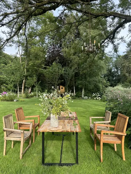Willeman, Fransa, 27 Temmuz 2024 Jardin du Rossignol Bahçeleri. Büyük bir ağacın altında ahşap sandalyeler ve masa seti olan güzel bir bahçe sahnesi. Ağaçtan sarkan dekoratif bir avize zarif bir dokunuş ekliyor. 