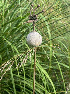 Willeman, France, July 27, 2024: Gardens Jardin du Rossignol. A unique garden stake with a bird-shaped head. Made of metal, stone, and plastic, it stands out against tall green grasses clipart