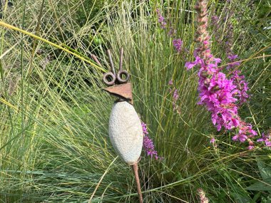 Willeman, France, July 27, 2024: Gardens Jardin du Rossignol. A unique garden stake with a bird-shaped head. Made of metal, stone, and plastic, it stands out against tall green grasses clipart