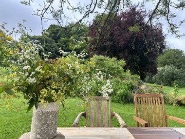 Dekoratif vazoda bir buket kır çiçeği. Canlı papatyalar, haşhaş tohumu kabukları ve bahçedeki ahşap bir masada dizilmiş yeşillik. 