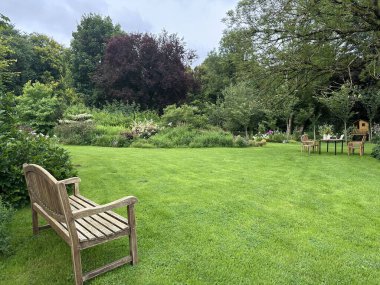 Ahşap banklı huzurlu bir bahçe. Uzayda yemyeşil bir çimen, yemyeşil bitkiler ve çiçeklerle çevrili, barışçıl bir geri çekilme yaratıyor. Jardin du Rossignol Bahçeleri, Fransa