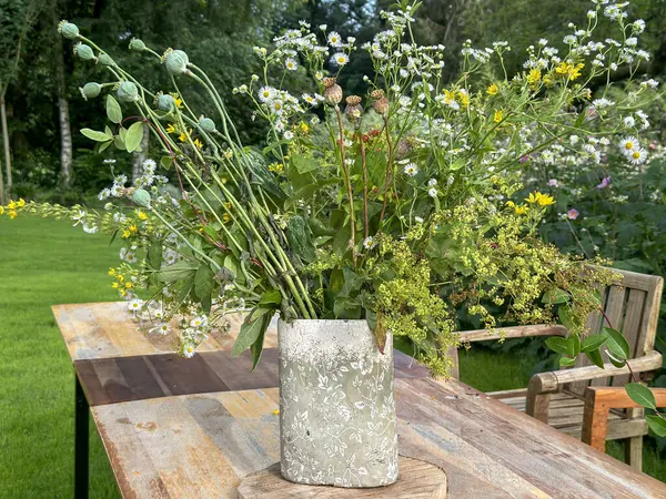 Dekoratif vazoda bir buket kır çiçeği. Düzenleme haşhaş tohumu kabukları, papatyalar ve yeşillik içeriyor. Bahçe düzeninde kırsal ahşap bir masa üzerine yerleştirilmiş. 'Jardin du Rossignol', Fransa