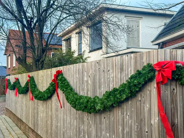 Sokakta noel süsü. Çitin üstünde kırmızı kurdeleler olan yeşil bir çelenk. Konut alanındaki ahşap bir çit boyunca süslü bir dış mekan dekorasyonu. Doğal ve canlı Noel dekoru olan tatil ruhu