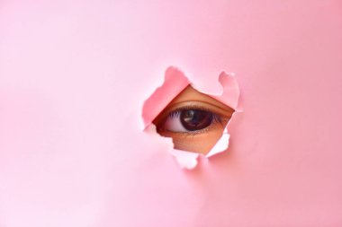 Pembe kağıt deliğinde küçük bir kızın gözünü gözlemlemek. Pembe kağıdın altındaki göz.