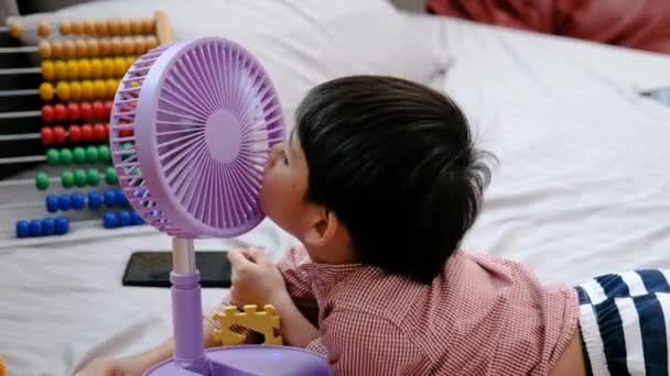 亚洲男孩在炎热的一天躺在床垫上快乐地玩着便携式风扇 — 图库视频影像