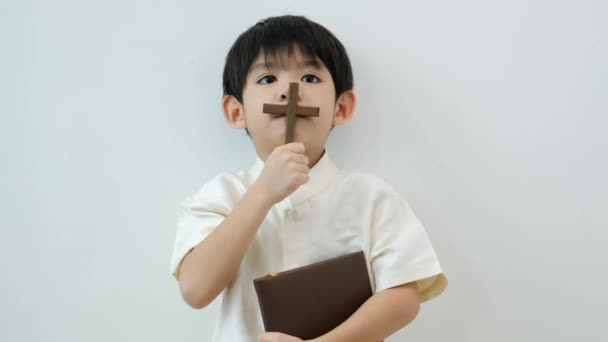 小さなアジアの少年は 十字架と宗教的な本 キリスト教の概念 4K決議を保持することを祈ります — ストック動画