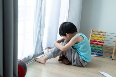 Üzgün Asyalı çocuk, başı öne eğik oturur ve kapıdan en sevdiği bebeğe bakar.