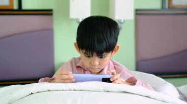 Küçük bir Asyalı çocuk yatağa uzanıp telefonuyla oynuyor..