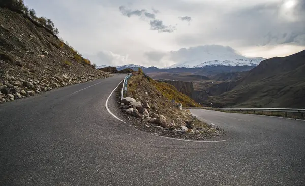 stock image The asphalt serpentine track winds and turns along the mountain slope against the backdrop of a mountain range with snow and glaciers, on a cloudy autumn day in the mountains