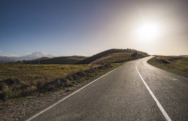 Bir dağ tepesinde asfalt yolda, akşam güneşinin ışığında, dağ sıralarının arasında yol al.