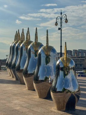 Yansıtıcı heykeller, kentsel bir alanda mavi gökyüzünün altında bir sırada dururlar..