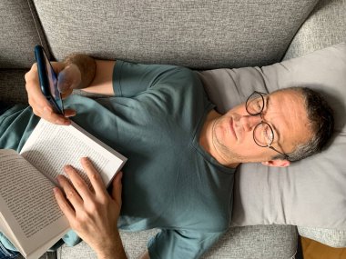 Orta yaşlı bir adam kanepede kitap okuyor. Orta yaşlı bir adam evdeki bir kanepede uzanıyor veya oturuyor, cep telefonu ve kitap okuyor. Evde sıcak bir hafta sonu. Kilitlenme. Kendini soyutlama. Tatilde...