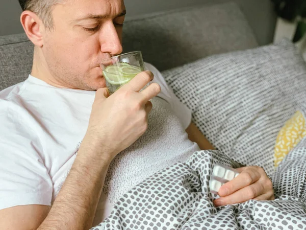 Middle aged man is sick at home, having flu or hangover. Middle aged man is in bed having flu covid or hangover, drinking water with lemon lime. Refreshing. Holding medical pills in hand. Holding