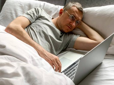 Middle aged adult man using laptop at home In bed vibes white bedsheets Alone Early morning working. freelancer, freelance job, using technology, using social media, watching movie, checking news