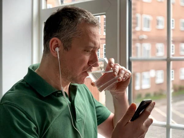 Middle aged man is talking on phone and drinking water. Video call. Middle aged man is talking on phone with friends or family, video call, watching video or checking news on social media. Staying at