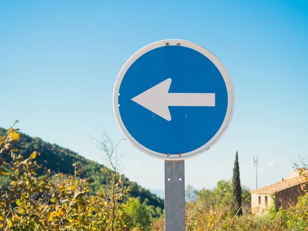 Close up of a turn left only direction sign with mountain on left side, a house on right side and a clear blue sky on a sunny day