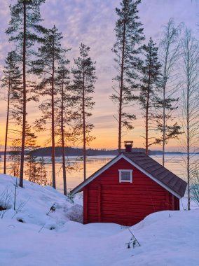 Finlandiya 'da günbatımı renklerinde göl kıyısına yakın ormanda kırmızı bir kulübe. 
