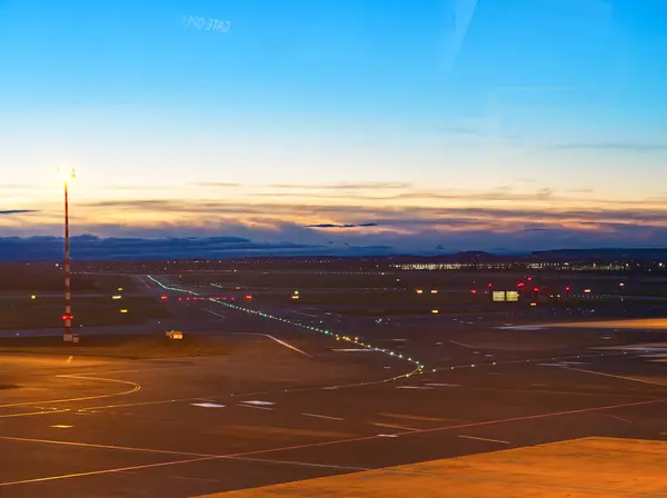 Aydınlatılmış havaalanı pisti alacakaranlıkta renkli gökyüzü ve uzak şehir ışıklarıyla