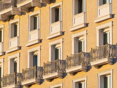 Süslü, demirden balkonları ve beyaz pencereleri olan sarı bir apartmanın ön cephesi.